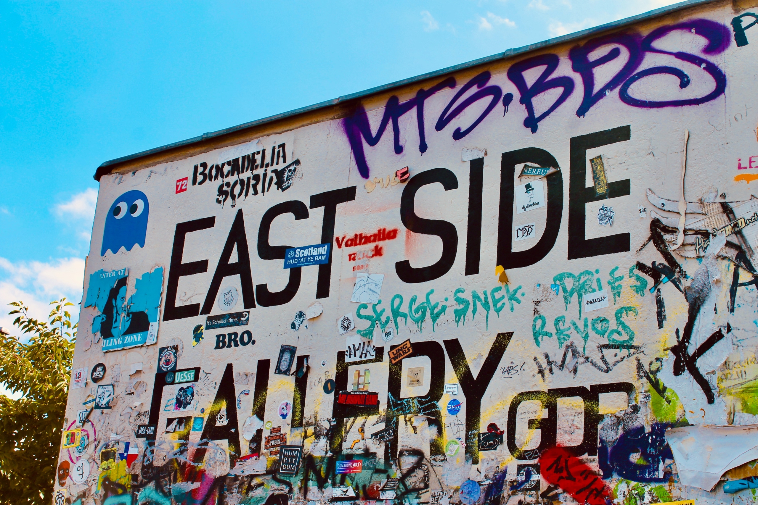 Graffiti-covered wall with bold "EAST SIDE GALLERY" text, colorful tags, stickers, and artwork, conveying urban creativity and vibrancy under a blue sky.