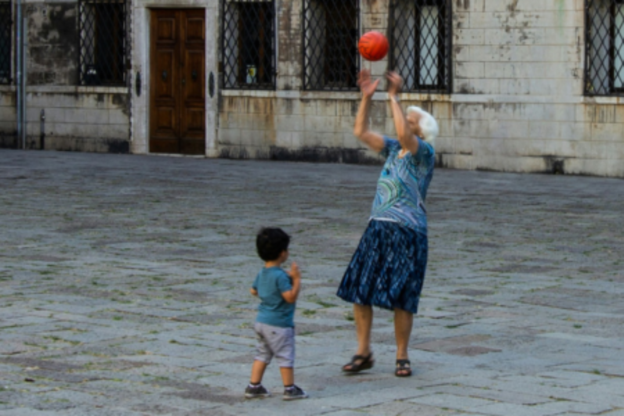 Elderly woman in a blue skirt tosses an orange basketball, and a young boy watches in a stone courtyard.