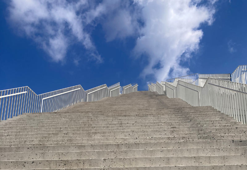 Concrete stairs lead upward to a bright blue sky with fluffy clouds. White railings line the stairs, creating a sense of openness and aspiration.