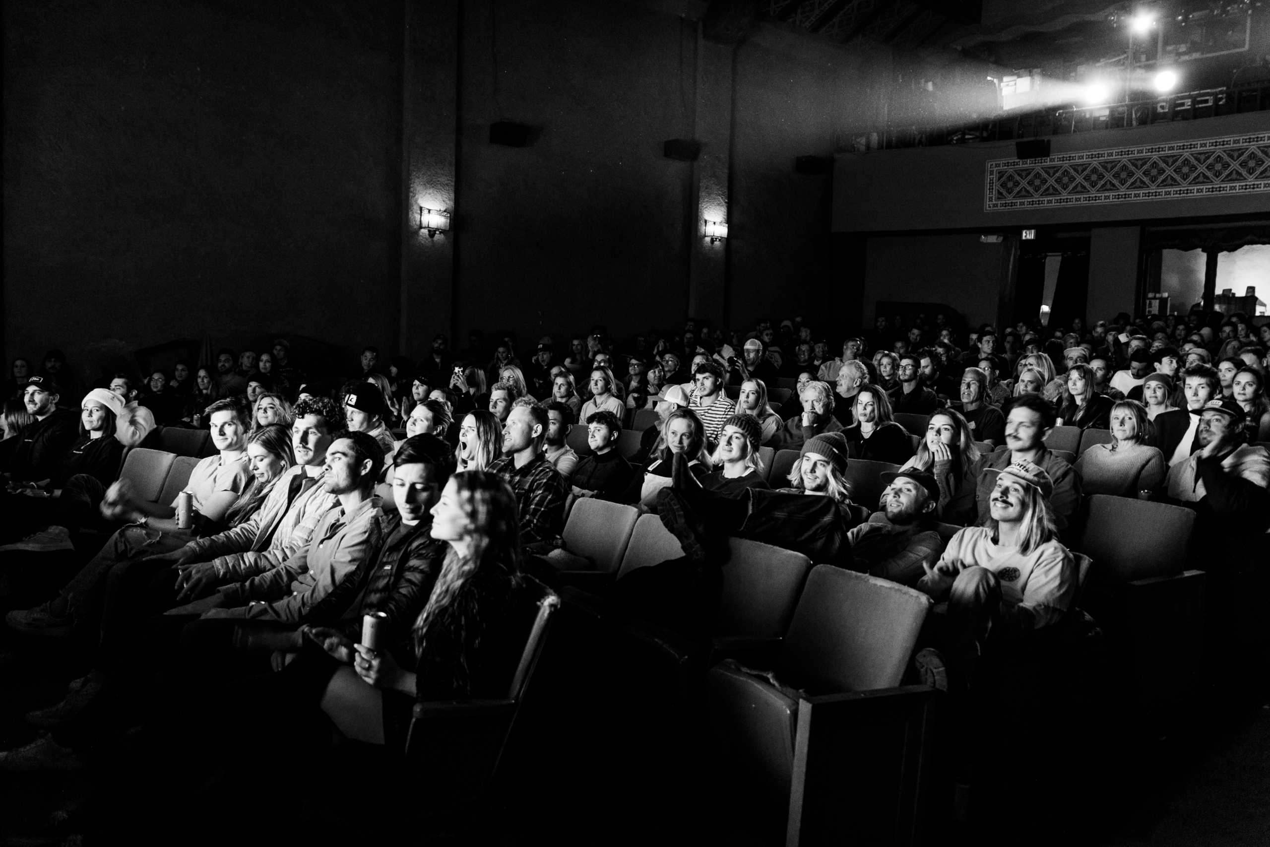Black and white photo of a packed theater audience smiling and focused on a screen. The atmosphere is lively and engaged, with soft ambient lighting.
