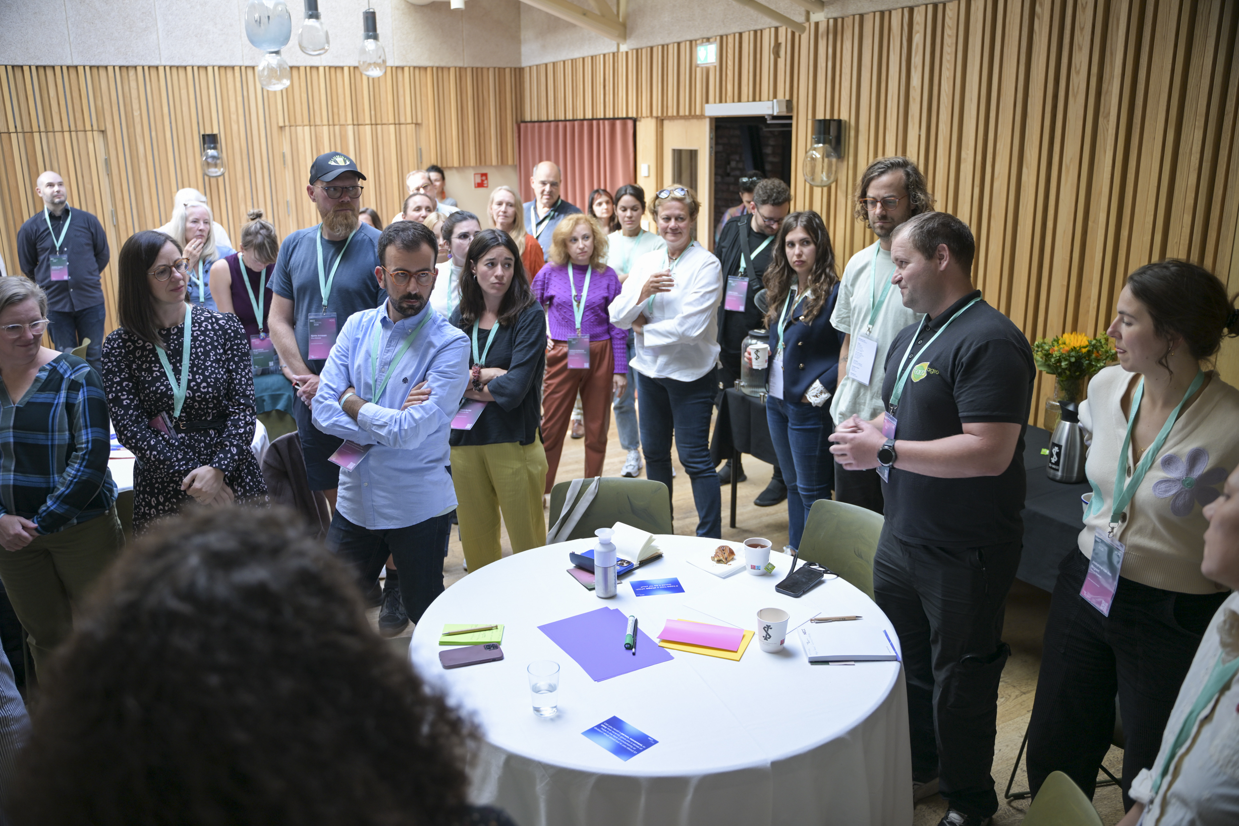 A diverse group of people stands attentively in a conference room, engaged in a discussion around a table with papers and pens during NECE Festival: HOPE.