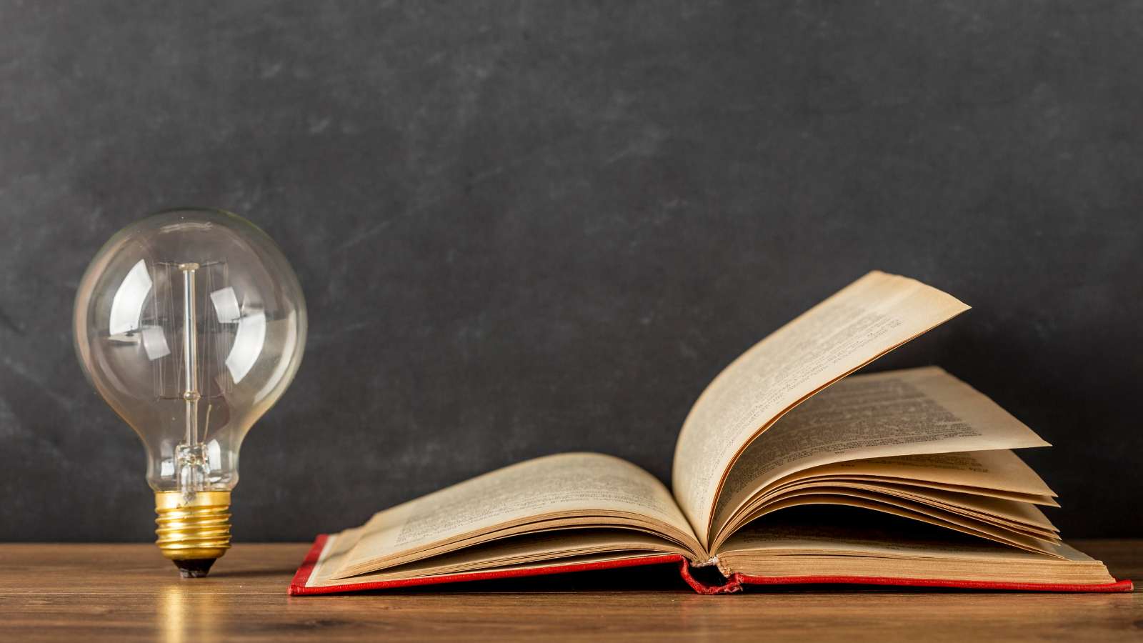 An open book with a red cover lies on a wooden surface next to a large, clear light bulb against a dark background, symbolizing knowledge and ideas.